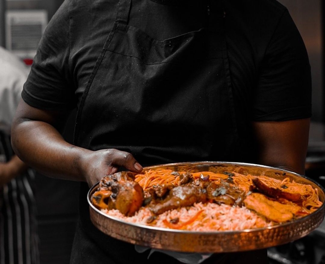 Chef holding a large platter with rice, pasta, meat, and sauce in a kitchen setting.